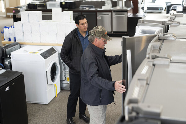 Two men in overcoats stand in an appliance showroom opening refrigerator doors to inspect them as one of the men shops.